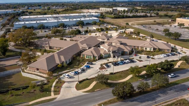 Aerial view of Sedona Trace Health & Wellness facility showing a large, single-story building with multiple peaked roofs, surrounded by parking lots, roads, and landscaped areas with trees and grass.