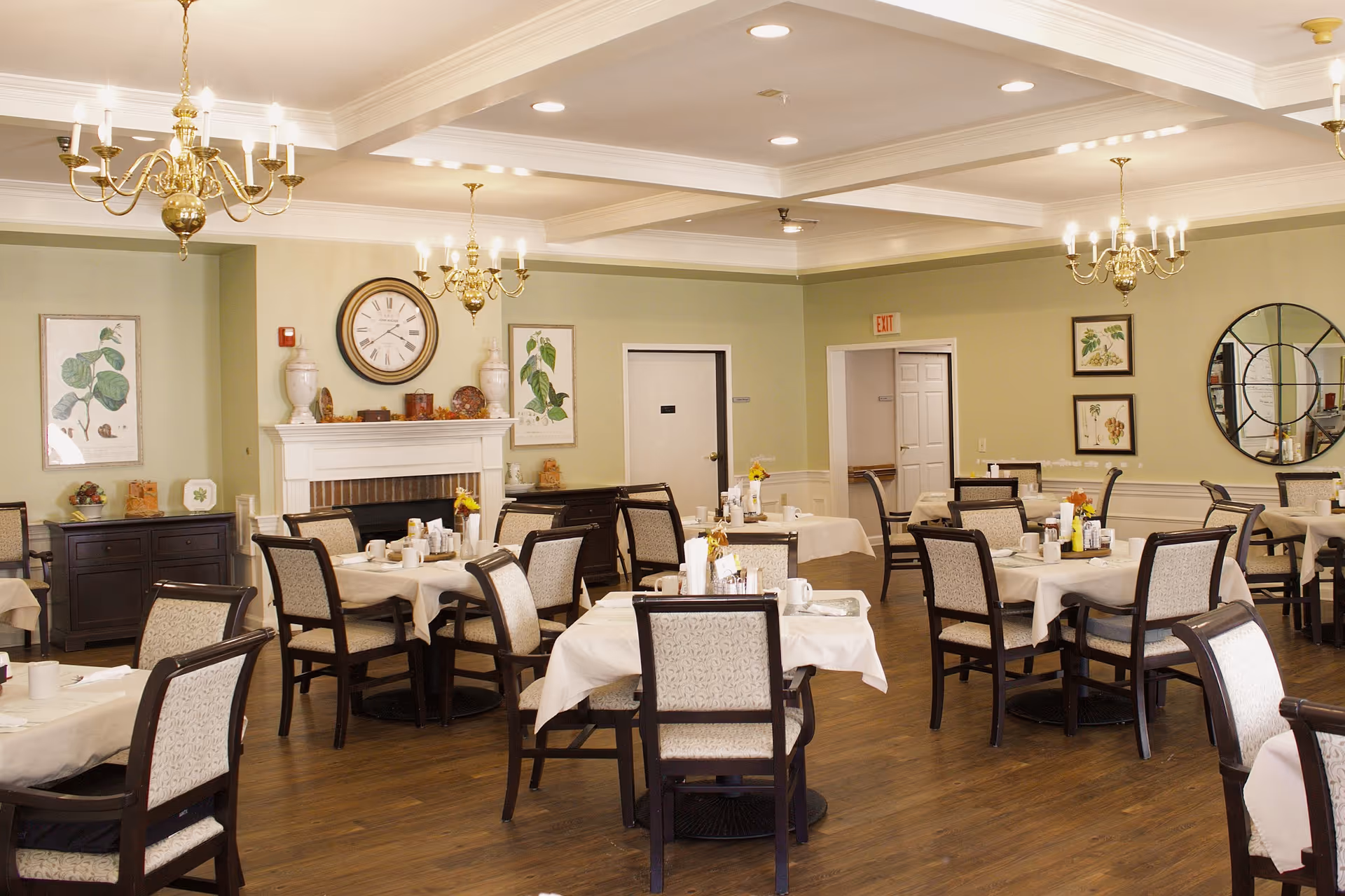 A well-lit dining room with multiple tables covered in white tablecloths, each set with cups, condiments, and small flower vases. The room features wooden chairs with cushioned seats and backs, a fireplace with decorative items and a large clock above it, light green walls adorned with botanical artwork, and three brass chandeliers hanging from a coffered ceiling.