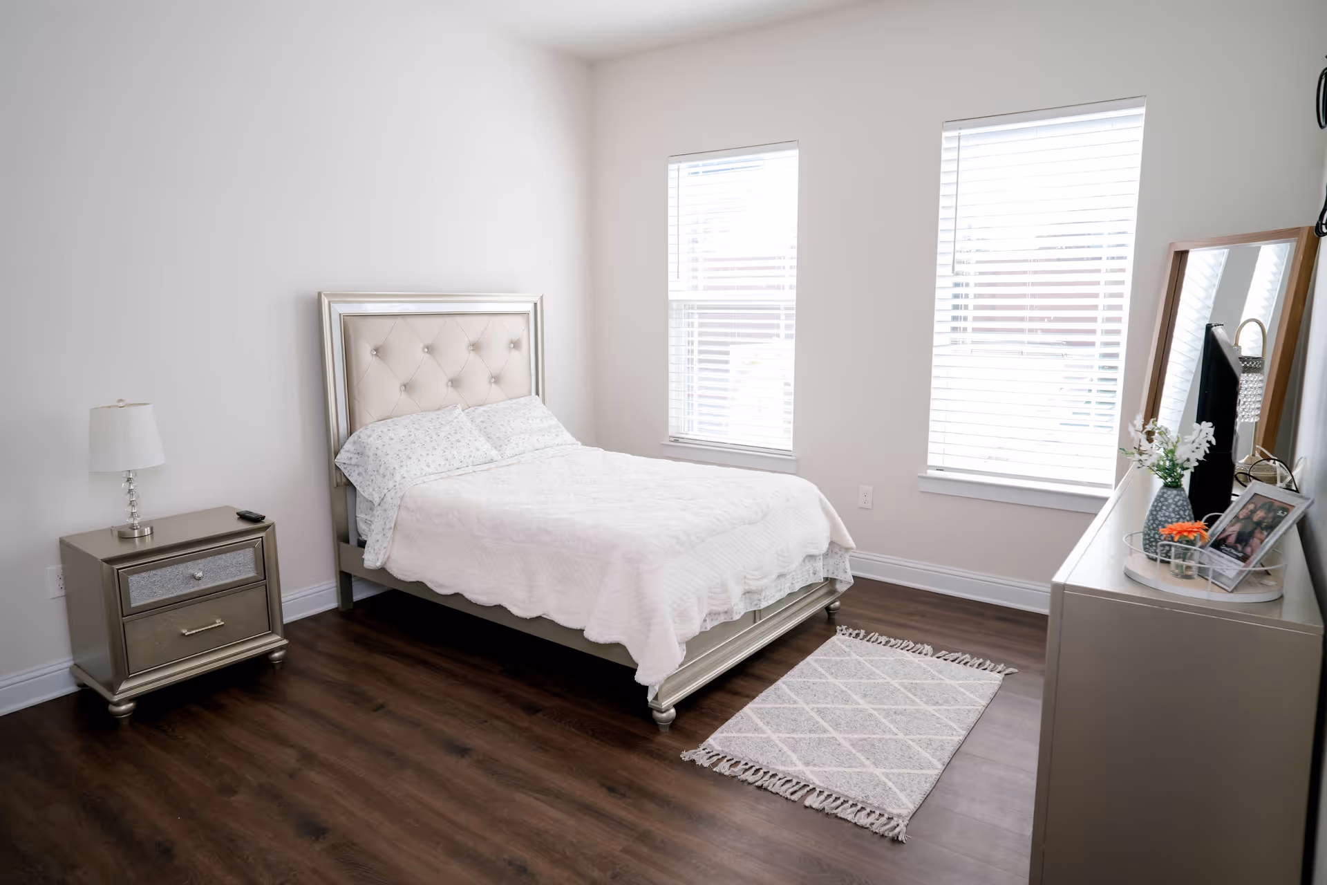 A bright and clean bedroom with a bed featuring a tufted headboard, white bedding, and two pillows. There is a nightstand with a lamp and a remote control on the left side of the bed. Two large windows with white blinds allow natural light to fill the room. On the right side, there is a dresser with a mirror, a vase with flowers, and a framed photo. A small patterned rug is placed on the wooden floor near the foot of the bed.