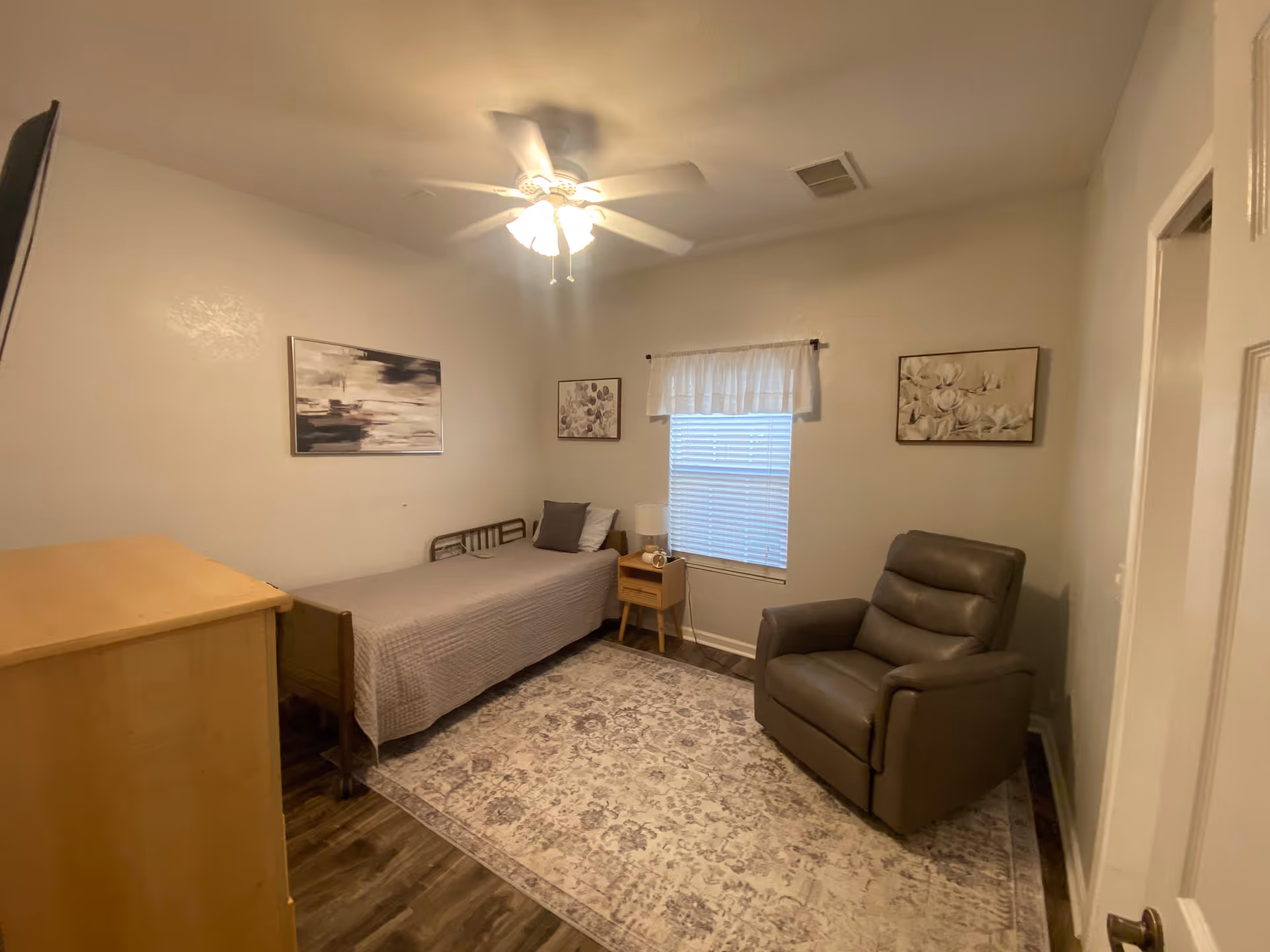 A small bedroom with a single bed covered in a gray quilt, a wooden dresser, a nightstand with a lamp and clock, a large patterned rug, a gray recliner chair, and three framed artworks on the walls. A ceiling fan with lights is on the ceiling, and a window with blinds and a white valance is centered on the far wall.