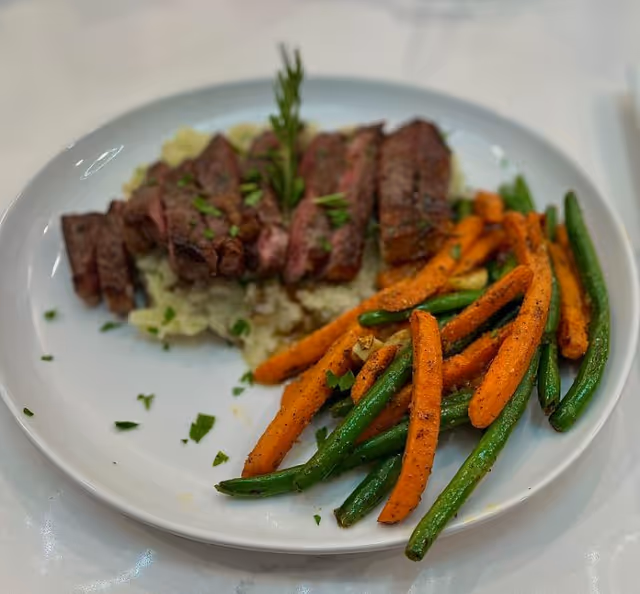 A plate of sliced steak on mashed potatoes served with roasted carrots and green beans, garnished with herbs.