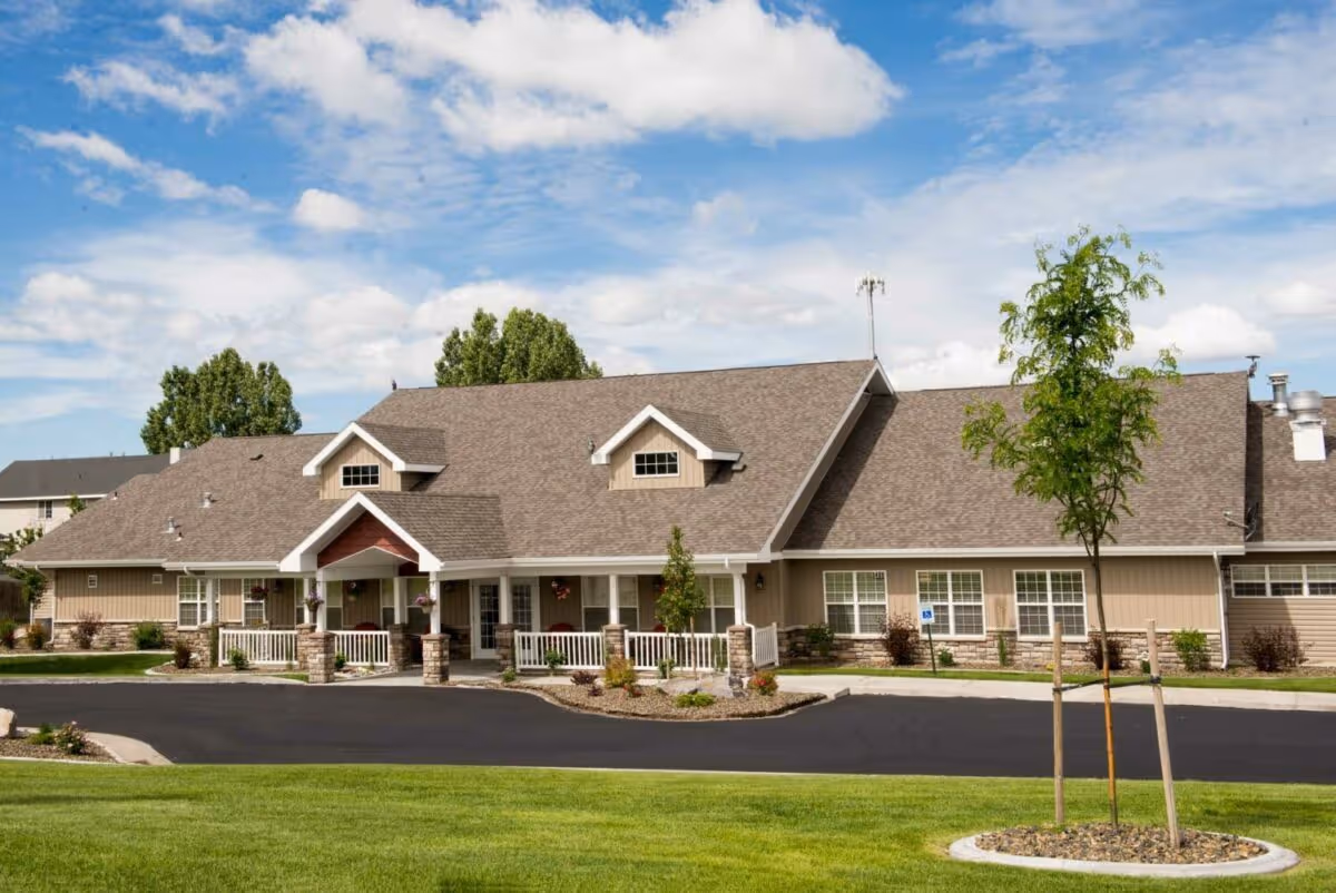 Exterior view of a single-story assisted living facility building with beige siding, a brown shingled roof, white trim, and a covered entrance supported by stone pillars. The building is surrounded by a well-maintained lawn, a paved driveway, and a few young trees under a partly cloudy sky.