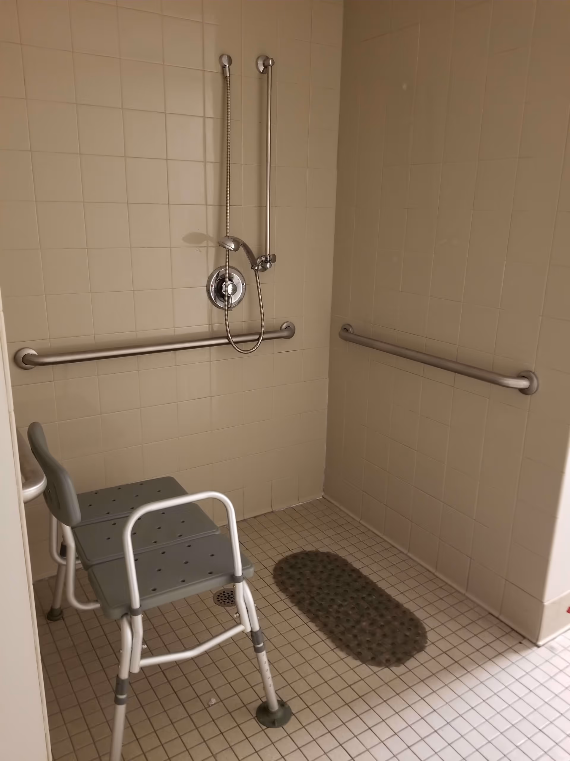Accessible shower area with beige tiled walls and floor, equipped with a handheld showerhead, multiple grab bars, a non-slip mat on the floor, and a gray shower chair with armrests and backrest.
