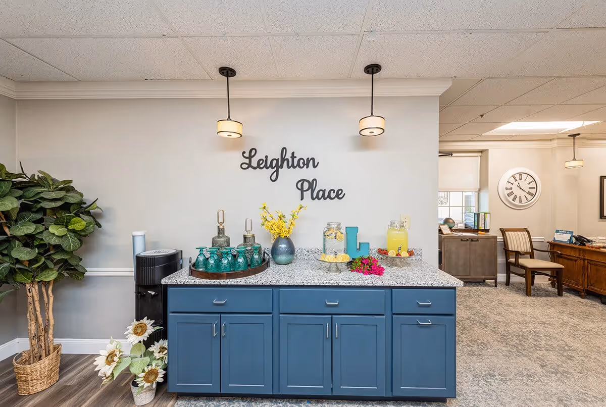 Interior view of a reception or common area at Leighton Place featuring a blue cabinet with a granite countertop. On the countertop are decorative items including a vase with yellow flowers, two glass dispensers with lemon water and lemonade, a tray with green glasses, and a blue letter L. Two pendant lights hang from the ceiling above the cabinet. To the left is a large potted plant and to the right is a seating area with chairs, a clock on the wall, and a wooden desk.