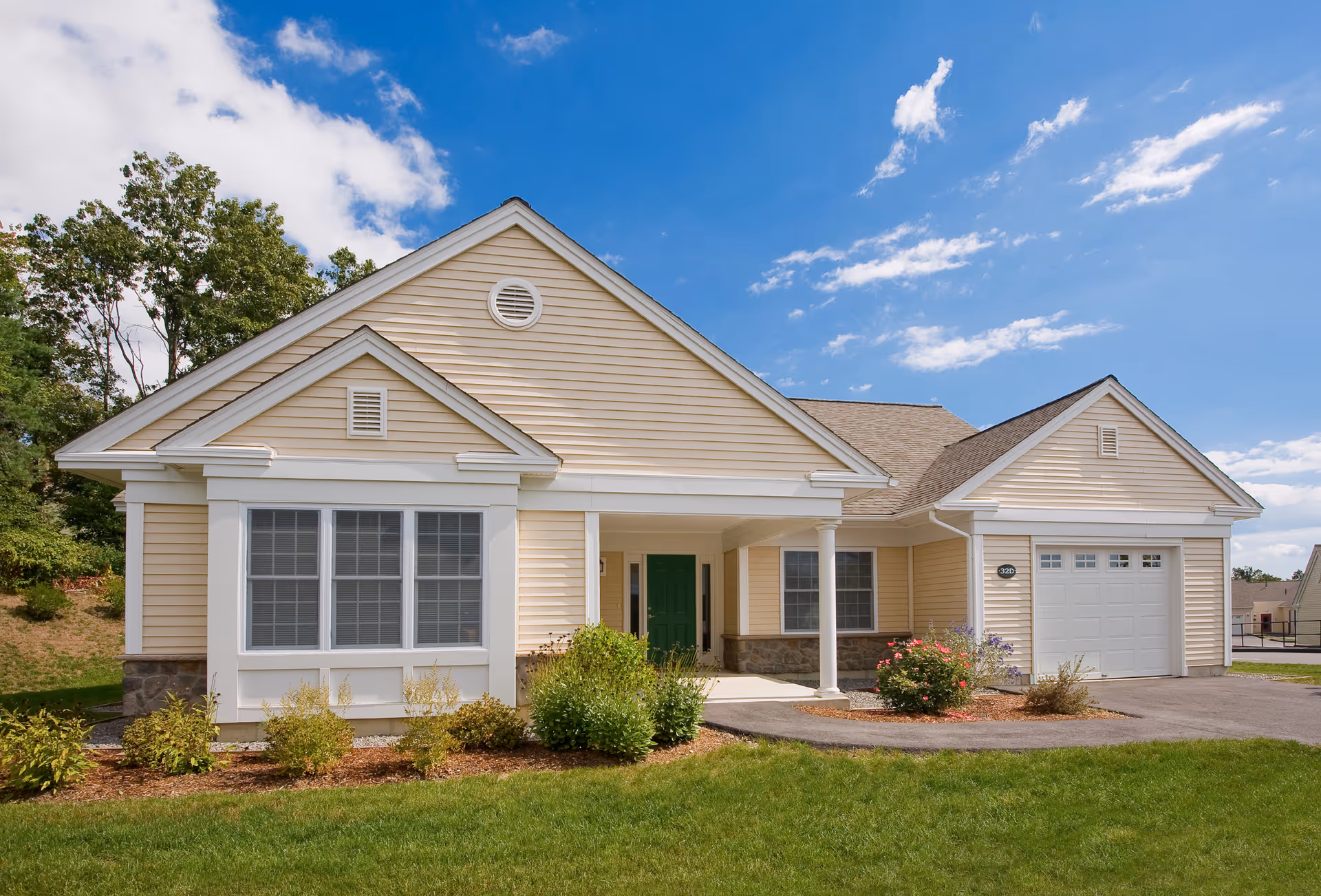Exterior view of a single-story beige house with white trim, a green front door, a garage, and a well-maintained lawn with shrubs and flowers under a partly cloudy blue sky.