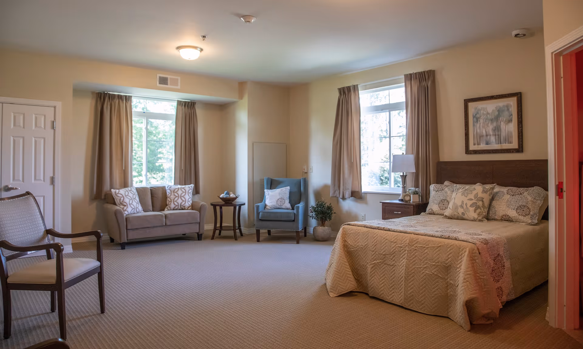 A spacious bedroom in The Ganzhorn Suites featuring a bed with floral bedding, a wooden nightstand with a lamp and clock, two windows with beige curtains, a blue armchair with a decorative pillow, a small round wooden table with a bowl, a beige loveseat with patterned pillows, and two wooden chairs. The walls are light-colored and there is a framed picture hanging above the bed.