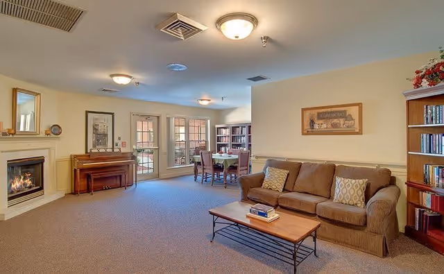 A cozy senior living common area featuring a brown sofa with patterned cushions, a wooden coffee table with books, a fireplace with a mirror above it, a piano, and a dining table with chairs near large windows letting in natural light. Bookshelves line the walls, and the room has a warm, inviting atmosphere.