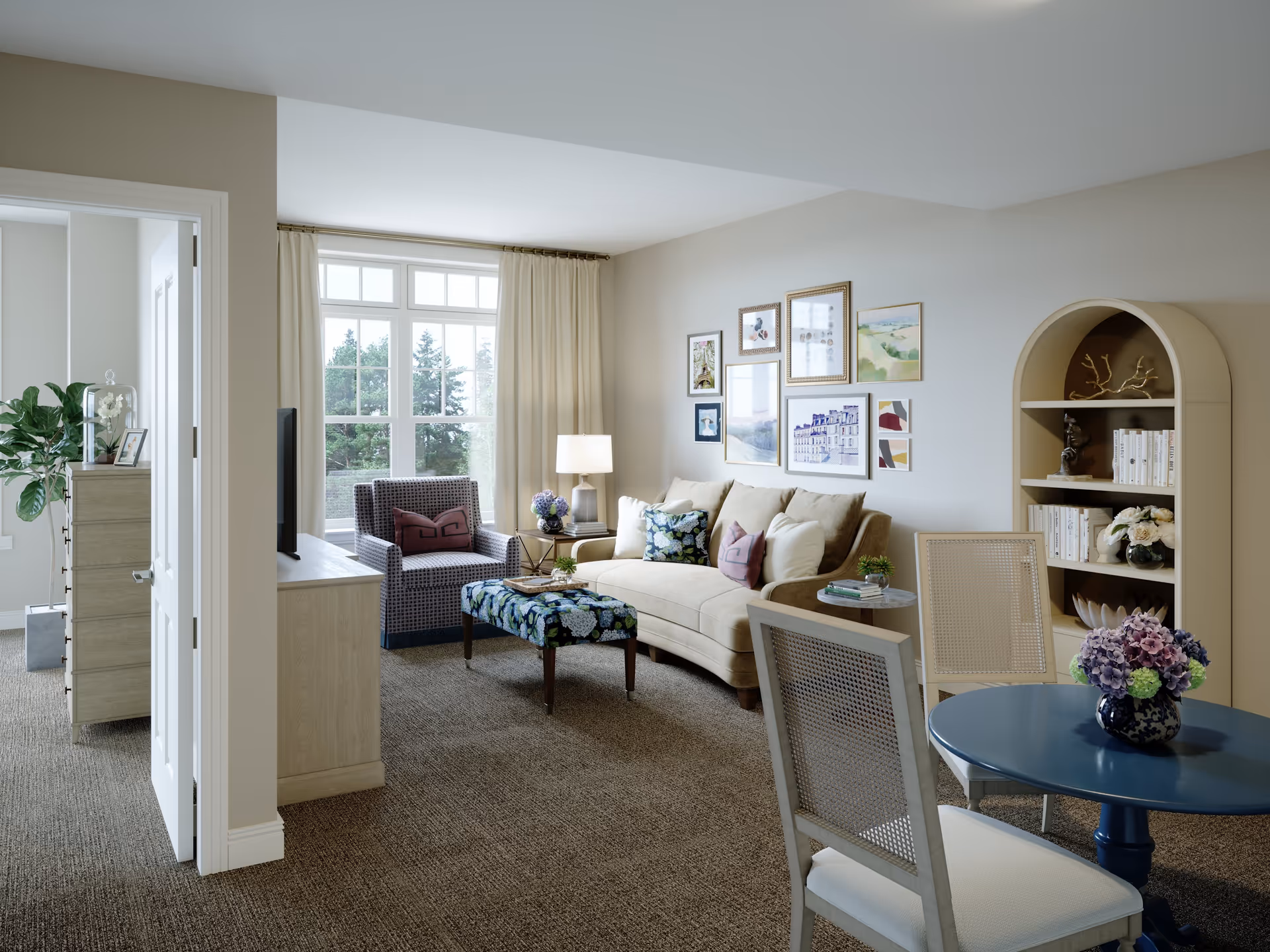 A cozy living room in a senior living facility with a beige sofa adorned with decorative pillows, a patterned armchair, a floral ottoman, and a blue round table with two chairs. The room features a large window with cream curtains, a bookshelf with books and decorative items, and framed artwork on the wall.