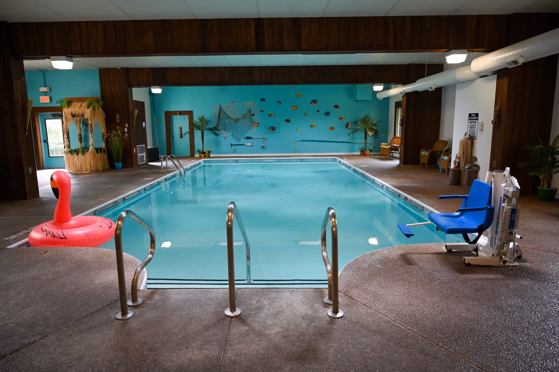 Indoor swimming pool with metal handrails, a blue pool lift chair on the right side, and a pink flamingo float on the left side. The pool area has turquoise walls decorated with colorful fish and nautical-themed decorations. There are chairs and plants around the pool area.