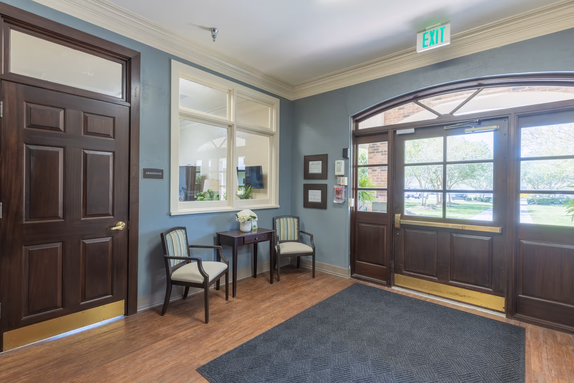 Entrance area of The Oaks of Houma facility with a wooden door labeled Admissions, two chairs with striped cushions, a small table with a flower vase, large windows, and a glass exit door showing greenery outside.