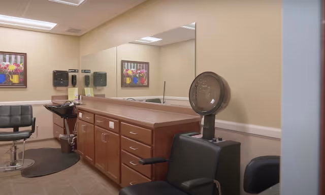 Interior view of a hair salon area in a senior living facility featuring a black salon chair with a hair dryer hood, a black styling chair, a countertop with multiple drawers and cabinets, a large mirror on the wall, two wall-mounted soap or sanitizer dispensers, and a framed floral painting.
