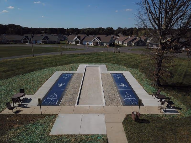 Aerial view of two outdoor shuffleboard courts with seating on a grassy area and residential cottages in the background.