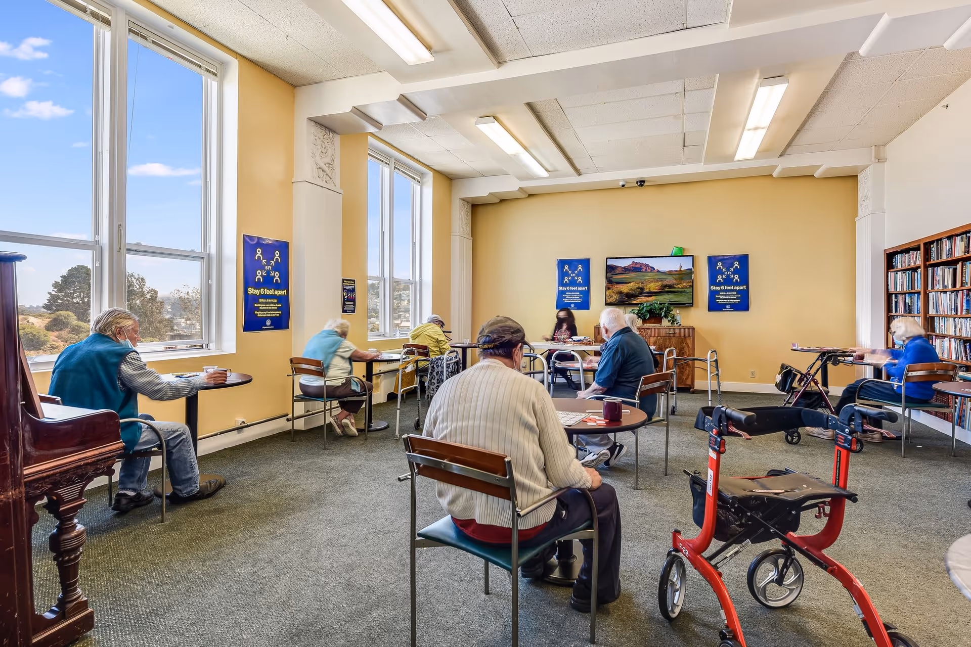 A group of elderly people sitting at separate tables in a spacious, well-lit room with large windows. The room has yellow walls, a bookshelf filled with books, and posters on the walls reminding people to stay 6 feet apart. A red walker is visible in the foreground.