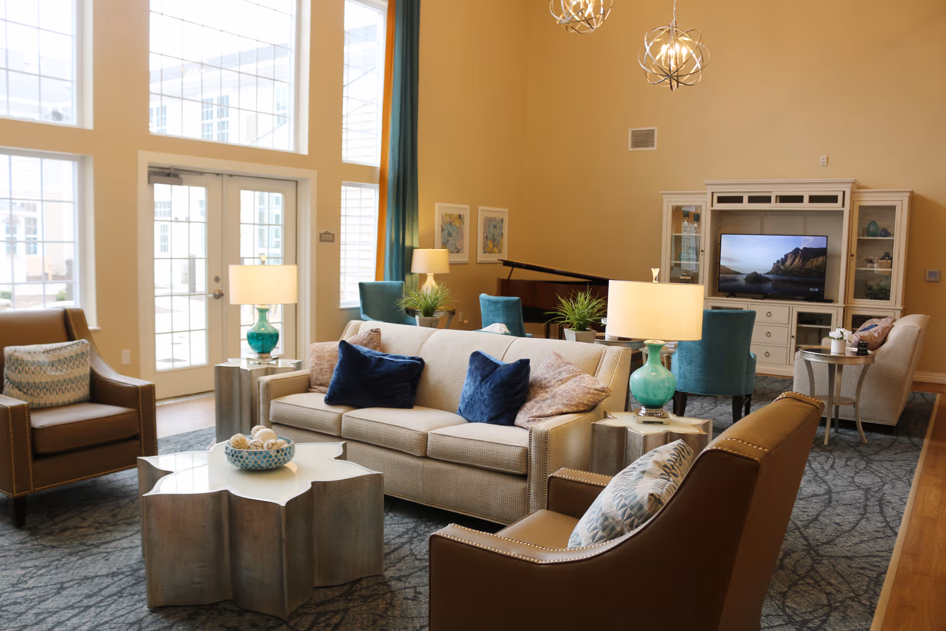 A bright and spacious living room in an assisted living facility with large windows letting in natural light. The room features a beige sofa with blue and patterned pillows, two brown armchairs with patterned cushions, a unique silver coffee table with a decorative bowl, and two turquoise lamps on side tables. In the background, there is a TV on a white entertainment center, teal chairs around a table, a piano, and framed artwork on the walls.