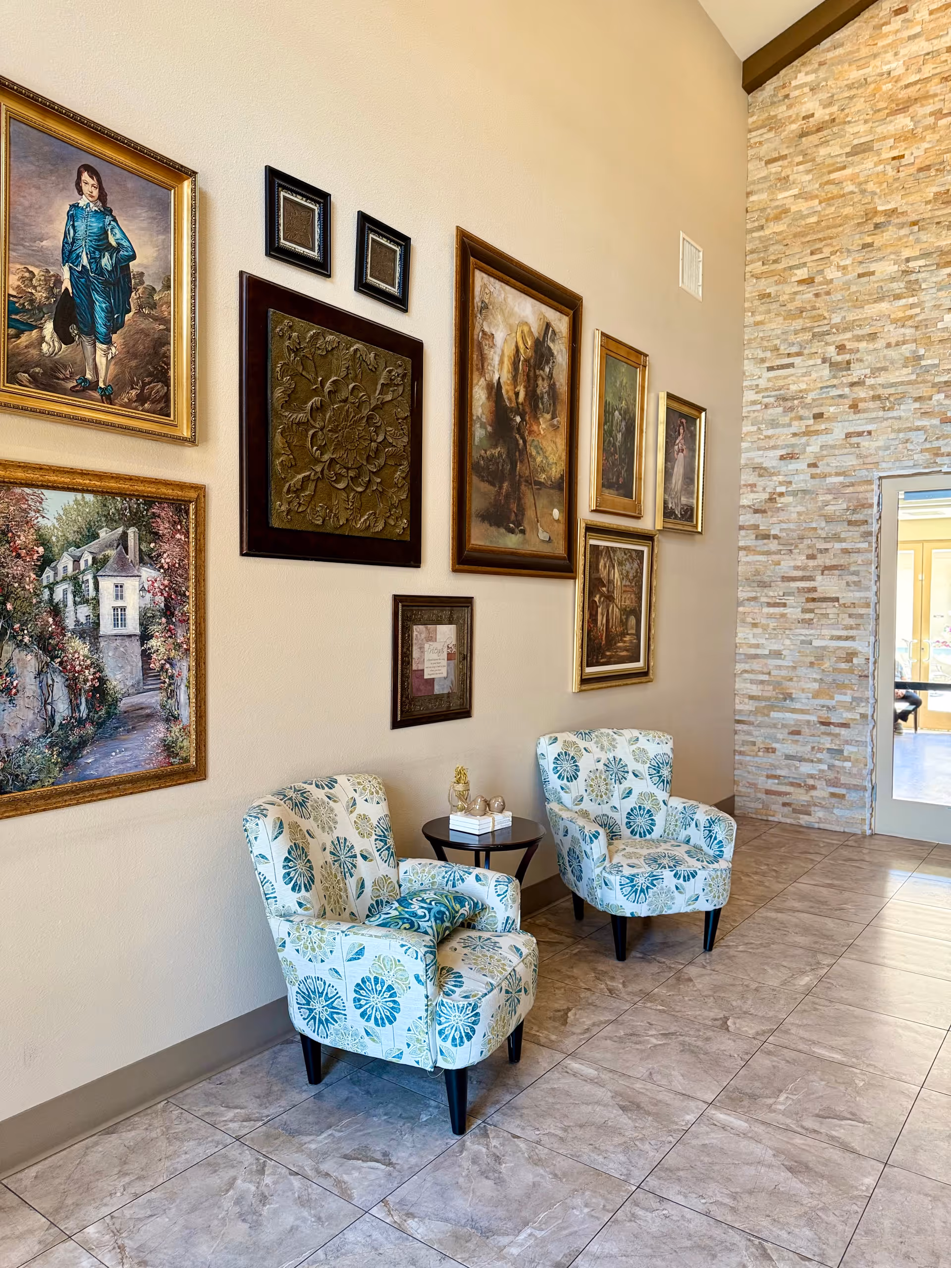 Two patterned armchairs and a small side table sit against a wall decorated with framed artwork and a stone accent wall in a tiled lobby.