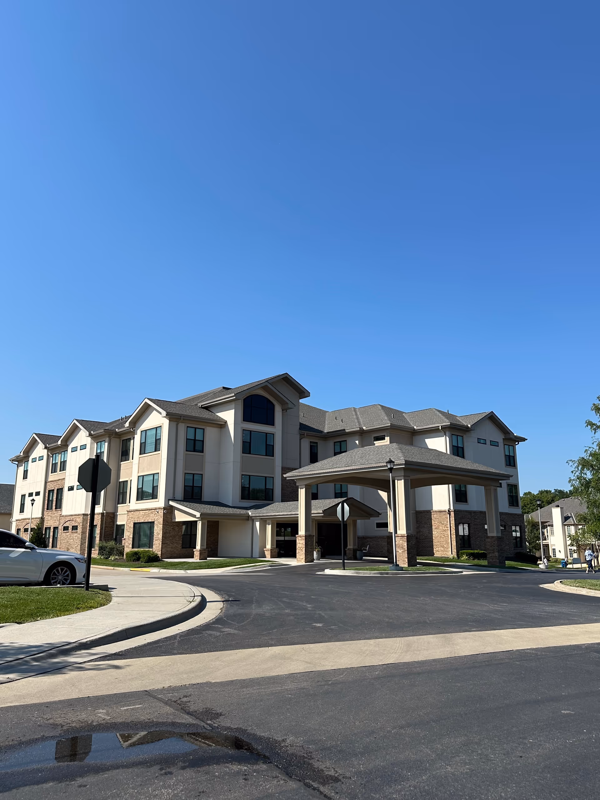 Exterior view of a three-story senior living facility building with a covered entrance driveway, surrounded by a paved road and some greenery under a clear blue sky.