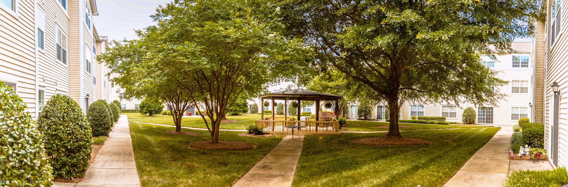 A landscaped courtyard area at The Dorchester facility featuring a central gazebo surrounded by green grass, trees, and shrubs. The courtyard is bordered by multi-story beige buildings with multiple windows and paved walkways leading to the gazebo.
