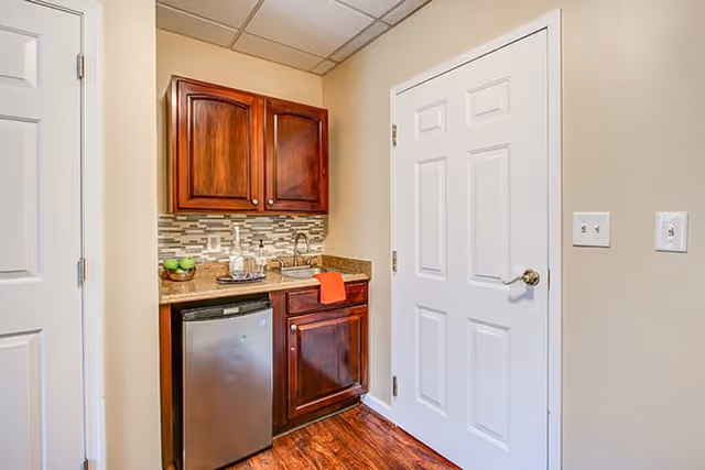 Small kitchenette with dark wood cabinets, a mini refrigerator, sink, and a white door.