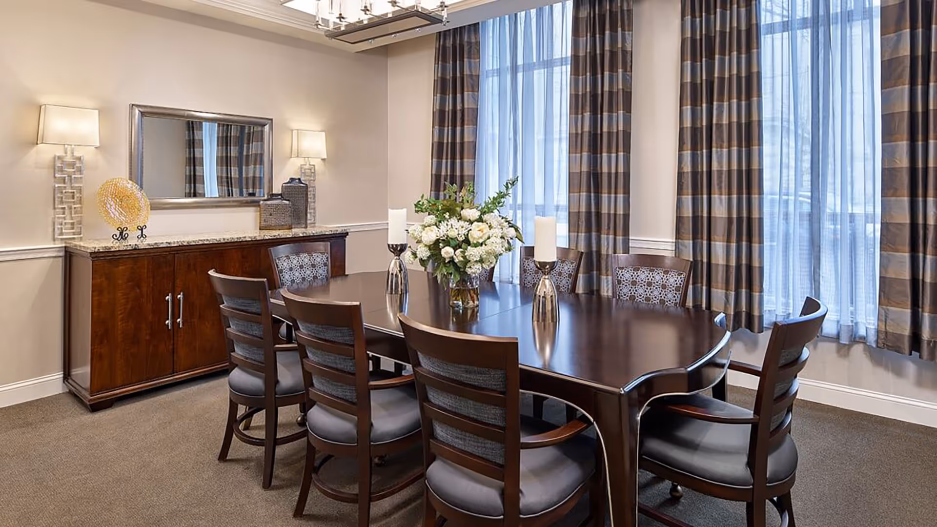 Elegant dining room with a dark wood table surrounded by chairs, a floral centerpiece, sideboard with mirror, and large windows with striped curtains.