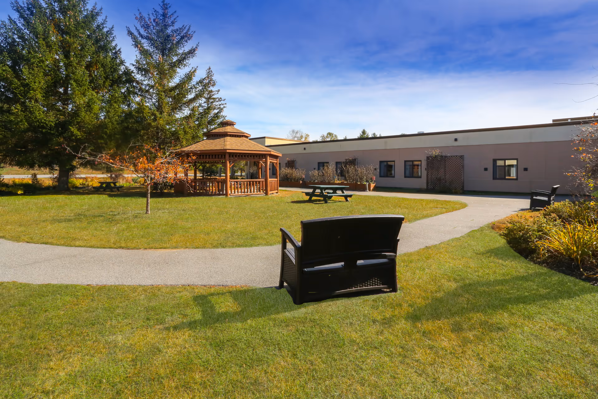 Outdoor garden area at Elderwood at North Creek featuring a wooden gazebo, picnic tables, benches, a paved walking path, and surrounding trees and shrubs under a blue sky.