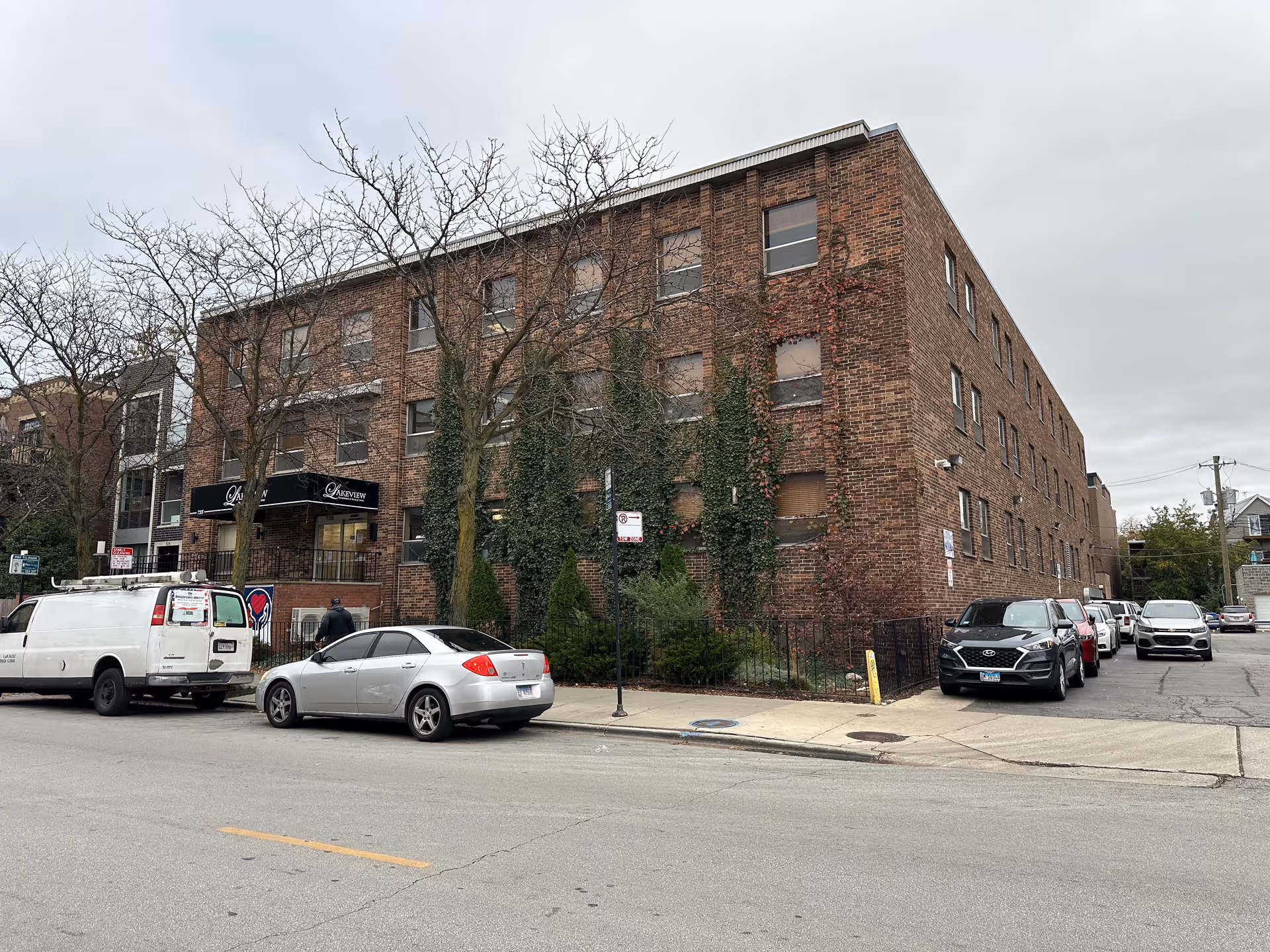 Exterior view of a three-story brick building with several windows, some covered with ivy and vines. There are leafless trees in front of the building and several parked cars along the street and driveway. A black awning above the entrance displays the name 'Lakeview'. The sky is overcast.
