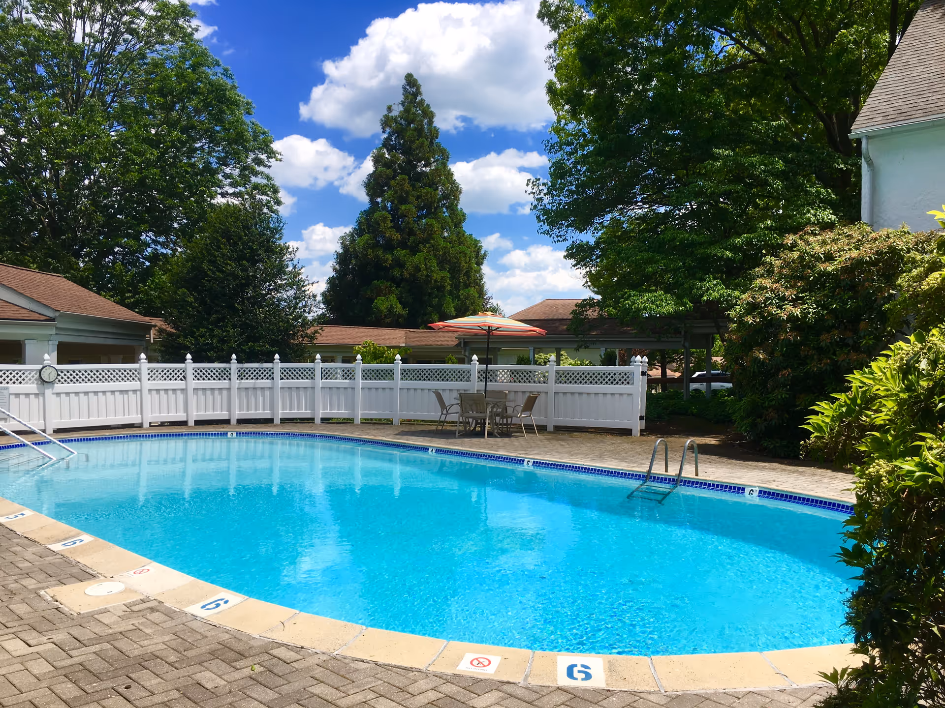 Outdoor swimming pool surrounded by a white fence with patio furniture and an umbrella nearby, lush green trees and bushes around, under a blue sky with scattered white clouds.