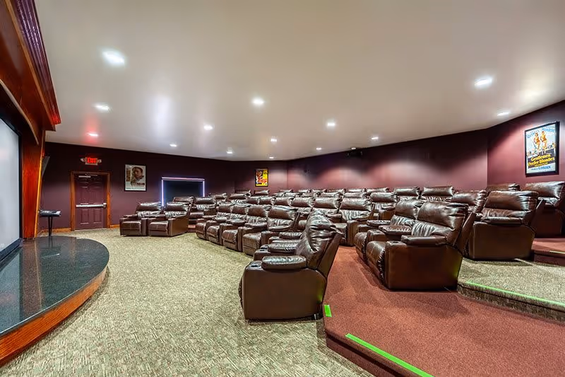 A spacious theater room with multiple rows of brown leather recliner chairs arranged in a tiered seating layout facing a large screen. The walls are painted dark purple and decorated with framed movie posters. The room is well-lit with recessed ceiling lights and has carpeted floors.
