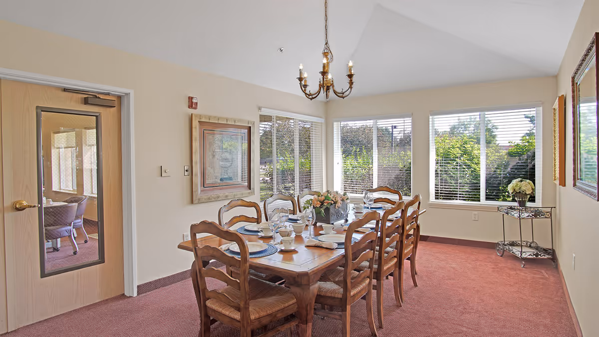 A dining room with a wooden table set for eight people, including plates, cups, glasses, and a floral centerpiece. The room has large windows with white blinds letting in natural light, beige walls, a red carpet, a chandelier hanging from the ceiling, framed artwork on the walls, and a small metal cart with a flower arrangement in the corner.