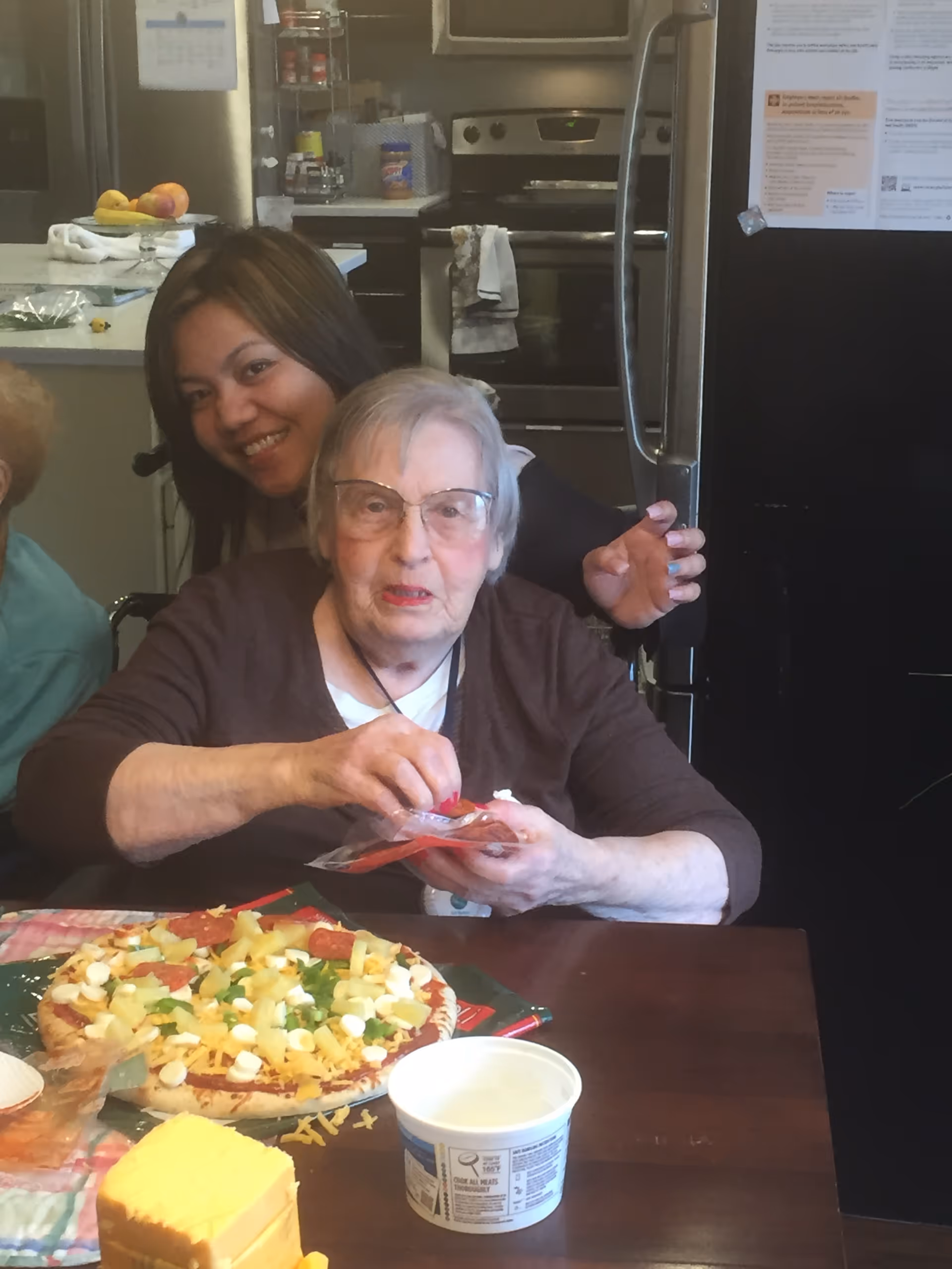 An elderly woman sits at a table assembling a pizza while a younger woman smiles behind her in a kitchen.