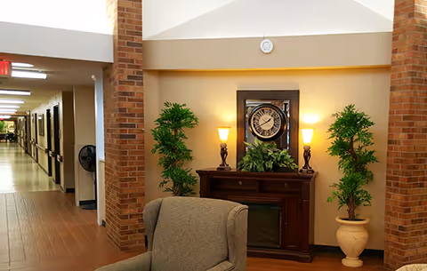 Interior view of a senior living facility hallway with a seating area in the foreground. The seating area includes a cushioned armchair, two potted plants, a wooden cabinet with a clock and two lamps on top. The hallway extends into the distance with doors and handrails along the walls.