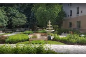 Outdoor garden area with a wooden bench in the center, surrounded by green shrubs and plants. A multi-tiered stone fountain is visible behind the bench. Trees and part of a building with windows are in the background.