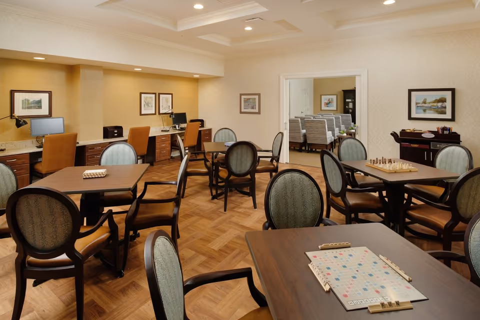 A well-lit common room with several tables and chairs arranged for games and activities. One table has a Scrabble board set up, and another has a chessboard. In the background, there is a row of desks with computers and chairs. The room has wood flooring, framed artwork on the walls, and a doorway leading to another room with additional seating.