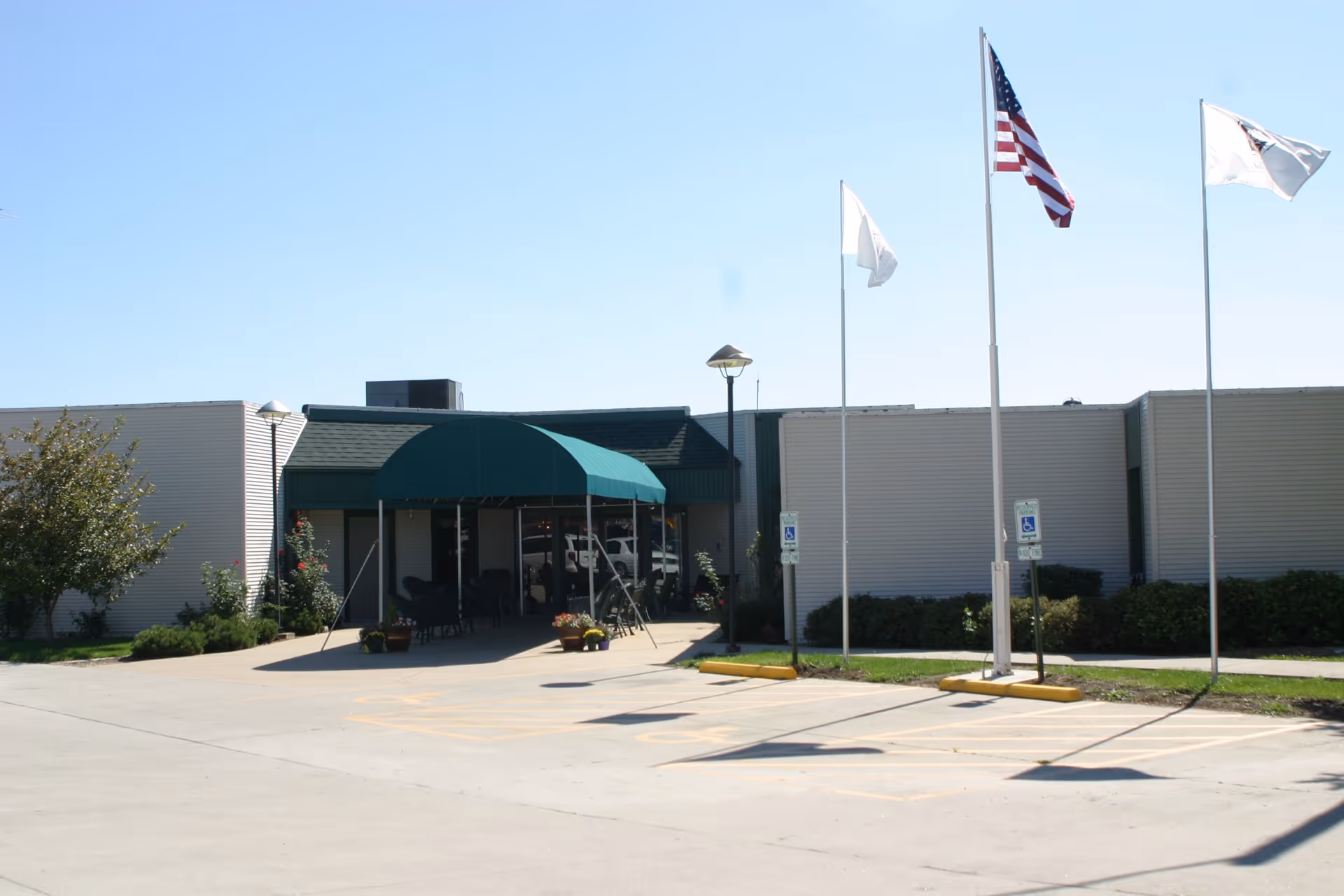 Exterior view of Sullivan Healthcare and Senior Living facility showing the entrance with a green awning, three flagpoles with flags including the American flag, and a parking area with handicap parking spaces.