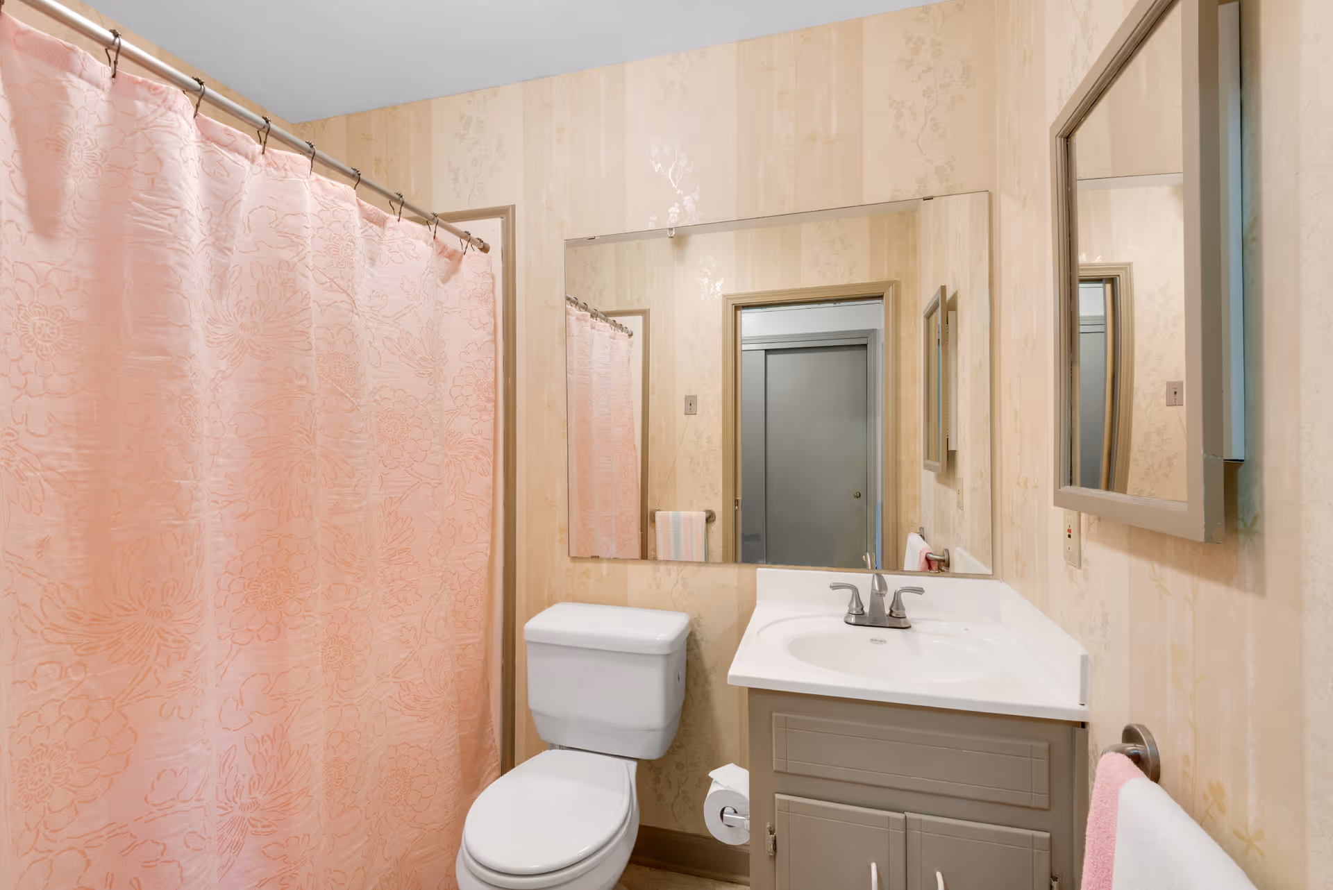 Bathroom with a pink floral shower curtain, toilet, sink vanity with mirror, and beige wallpaper.