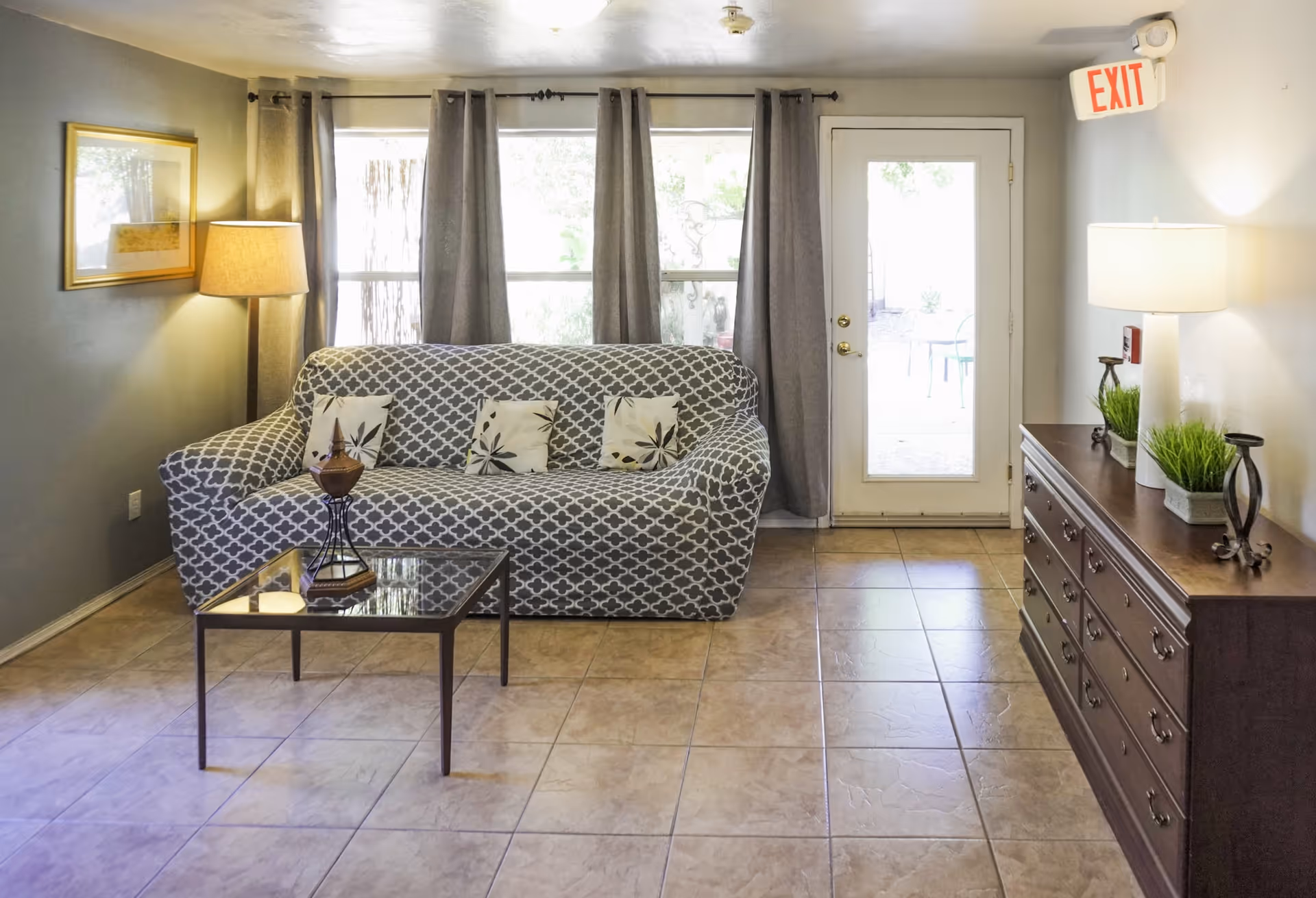 A cozy living room with a patterned gray sofa adorned with three decorative pillows, a glass coffee table with a small decorative item, a floor lamp beside the sofa, and a wooden dresser with two table lamps and green plants. The room has tiled flooring, gray curtains covering windows, and a glass door with an exit sign above it.