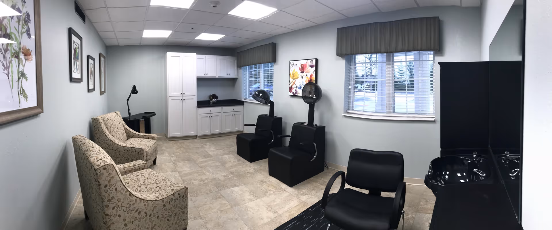 Interior of a hair salon area in a senior living facility with two beige patterned armchairs on the left, two black salon chairs with hair dryers in the middle, and a black salon chair with a sink on the right. The room has light gray walls, tiled floor, two windows with blinds, white cabinets, and framed floral artwork on the walls.