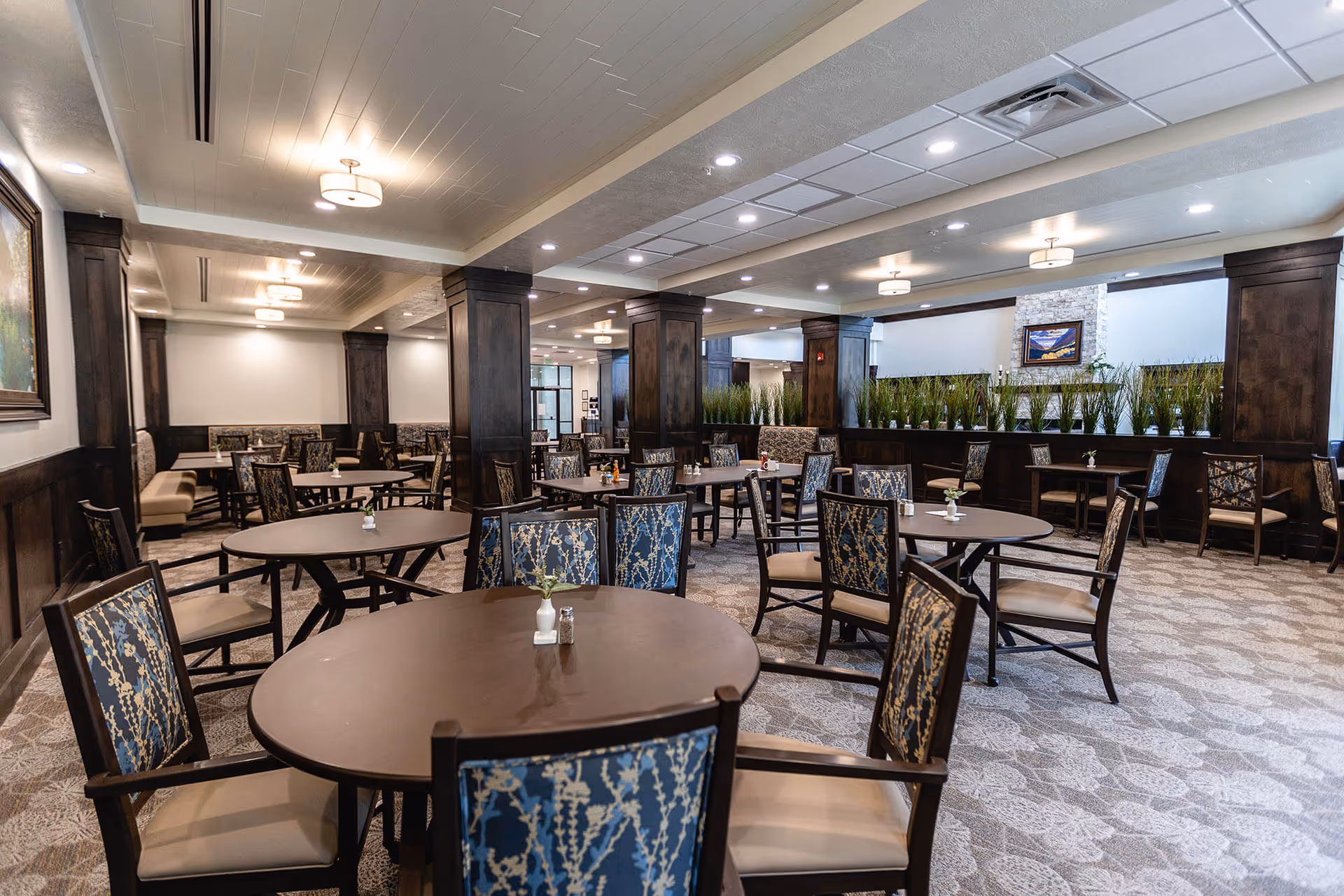 A spacious dining room with multiple round tables and chairs arranged neatly. The chairs have patterned upholstery with blue and beige tones. The room features dark wood paneling on the walls and columns, light-colored carpet with a subtle pattern, and ceiling lights providing warm illumination. There are decorative plants along a divider in the background and framed artwork on the walls.