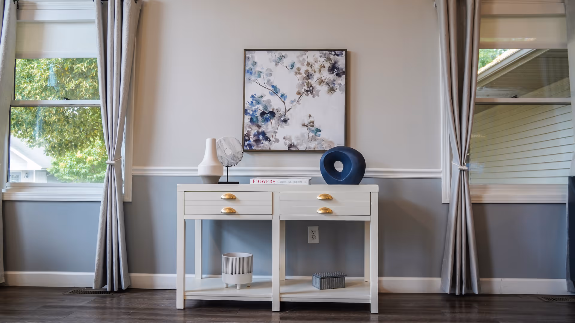 A bright interior room with two large windows on either side, each adorned with light gray curtains tied back. Between the windows is a white console table with gold handles, decorated with a white vase, a small fan, a blue circular sculpture, and a book titled 'FLOWERS'. Above the table hangs a framed abstract floral painting with blue and gray tones. The walls are painted white on the upper half and gray on the lower half, separated by a white chair rail. The floor is dark wood.