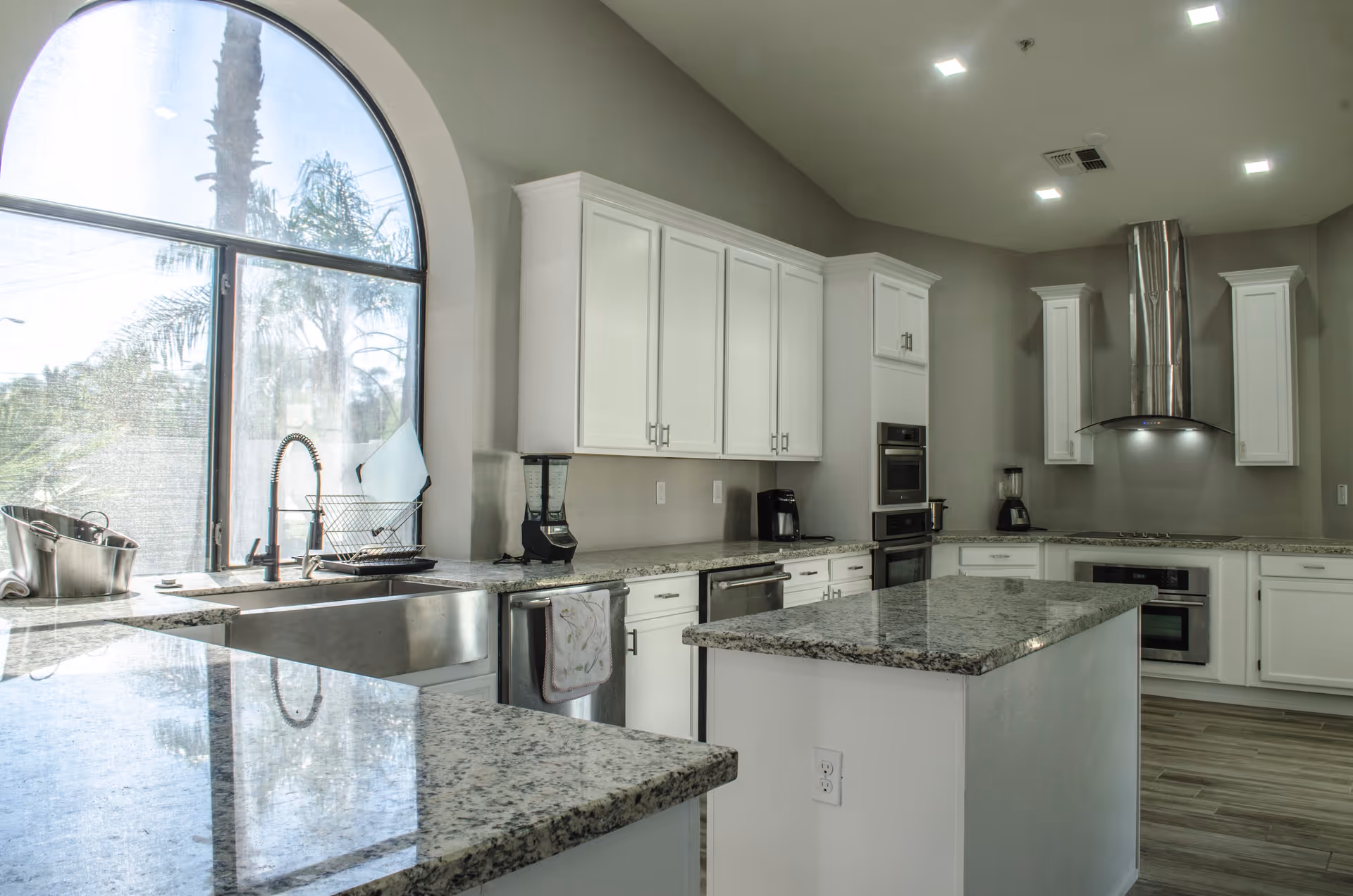 Bright modern kitchen with white cabinets, granite countertops, a large arched window, stainless steel sink, built-in oven, stovetop with a stainless steel range hood, and kitchen island.