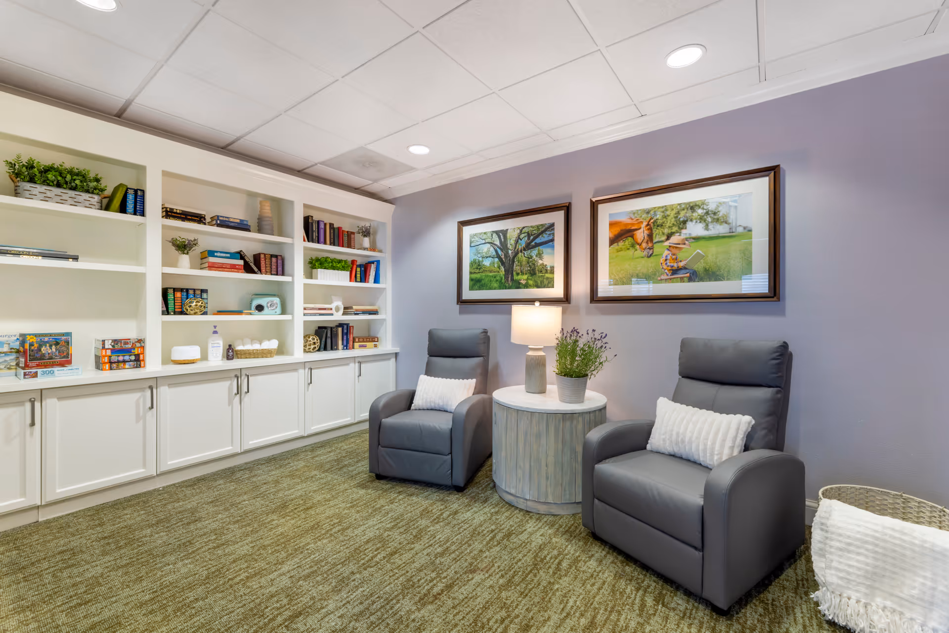 A cozy sitting area in a senior living facility with two gray armchairs, each with a white pillow, positioned around a round wooden side table with a lamp and a potted plant. Behind the chairs is a lavender-colored wall with two framed pictures, one of a tree and the other of a child reading with a horse. To the left, there is a built-in white bookshelf filled with books, plants, and decorative items. The floor is carpeted in a greenish tone.
