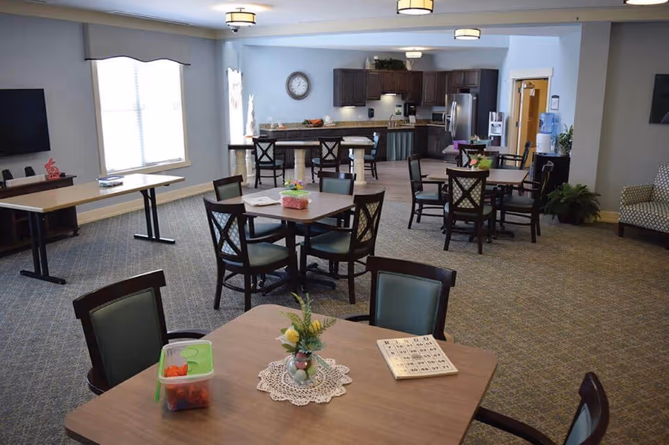 A spacious common area in a senior living facility featuring multiple tables and chairs arranged for group activities or dining. The room has a carpeted floor, large windows allowing natural light, a wall-mounted TV, and a kitchen area with dark wood cabinets and stainless steel appliances in the background. There are decorative plants and a bingo card on one of the tables, suggesting a recreational setting.