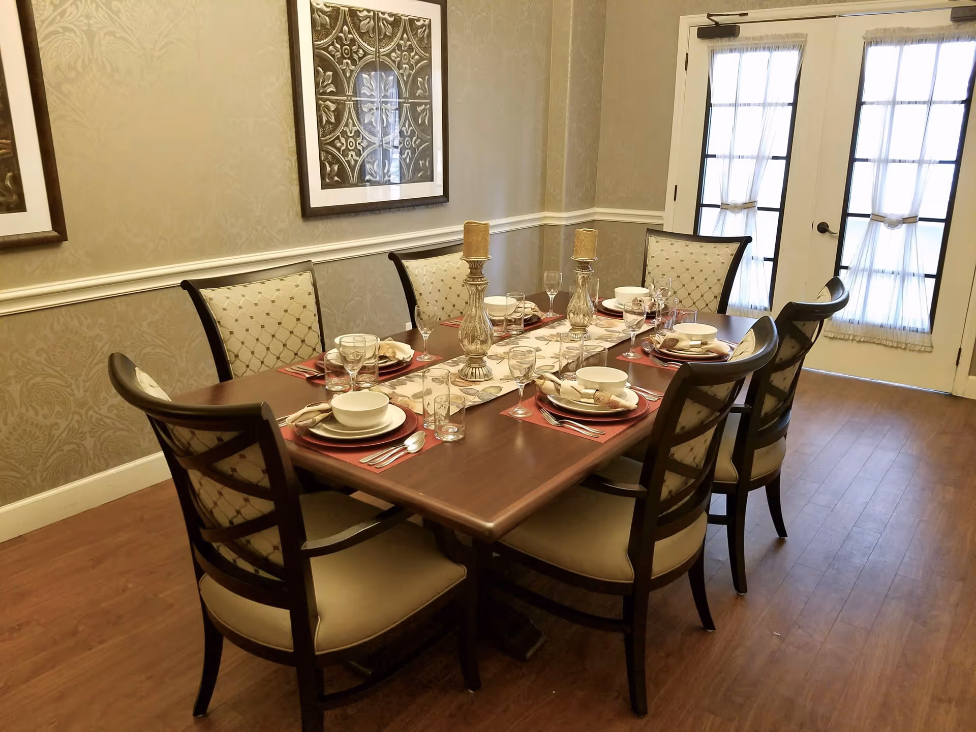 A formal dining room with a rectangular wooden table set for six people. The table is arranged with plates, bowls, glasses, silverware, and two decorative candle holders in the center. The chairs have cushioned seats and backs with a patterned fabric. The room has light-colored wallpaper with a subtle pattern, two framed artworks on the wall, and double doors with sheer curtains letting in natural light.
