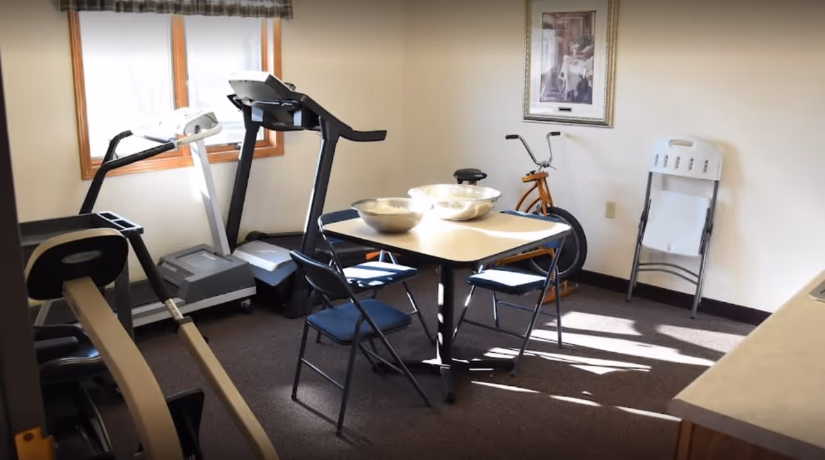 A small exercise room with a treadmill, stationary bike, and exercise equipment. In the center, there is a square table with four folding chairs around it and two metal bowls on top. A window lets in natural light, and a framed picture hangs on the wall. A folded chair leans against the wall near the exercise bike.