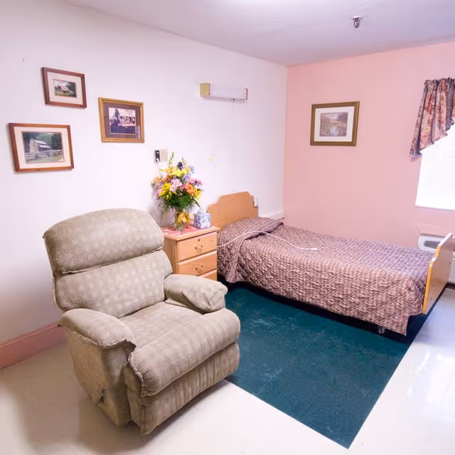 A small bedroom in a senior living facility with a single bed covered in a brown quilt, a wooden nightstand with a vase of colorful flowers, a beige recliner chair, and framed pictures on the walls. There is a window with floral curtains letting in natural light.