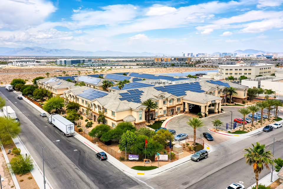 Aerial view of a senior living facility with solar panels on the roof, surrounded by roads, palm trees, and parked cars. The building is situated in a desert-like area with mountains in the background and a city skyline visible in the distance under a partly cloudy sky.