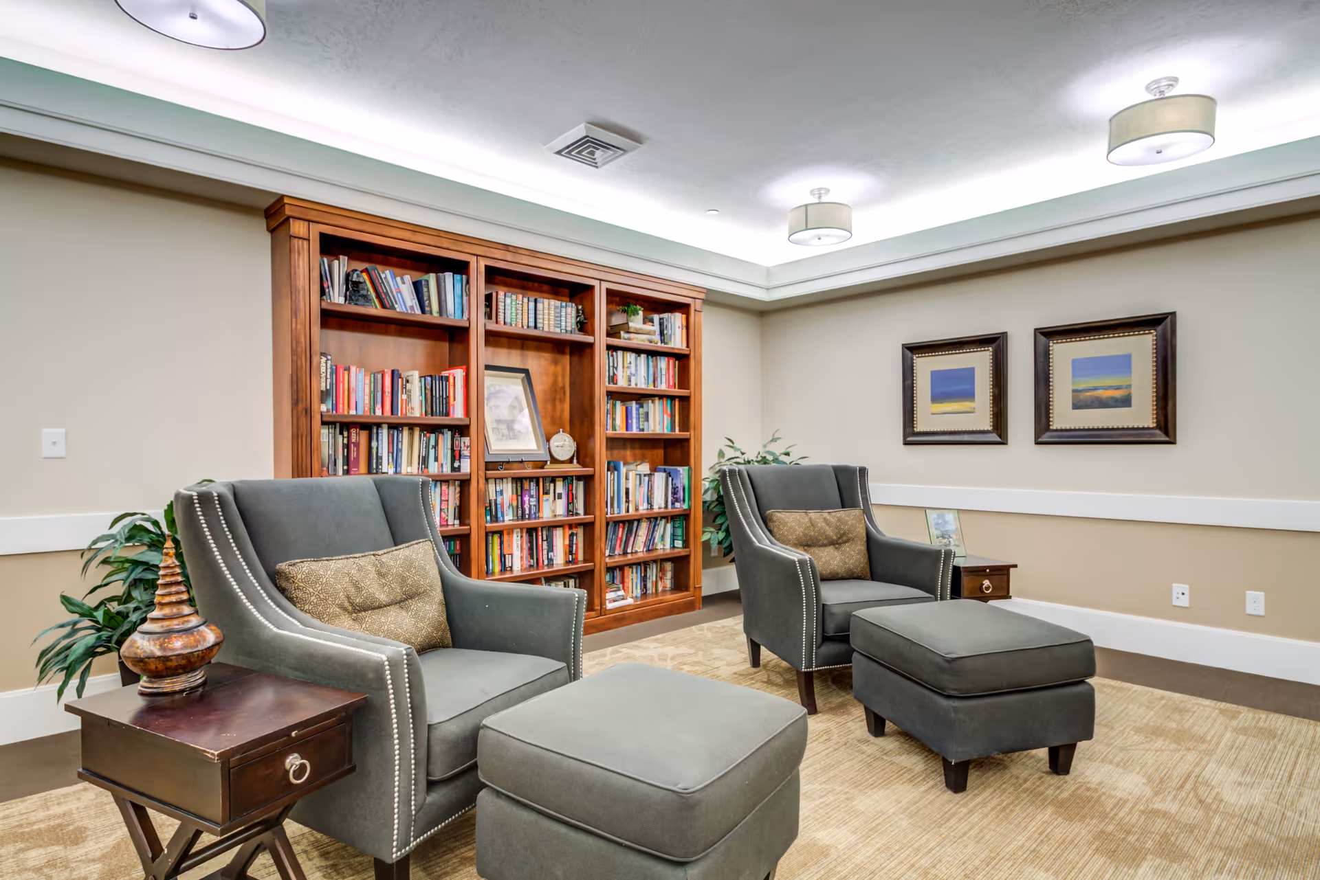 A cozy senior living common area with two gray armchairs each paired with a matching ottoman. Between the chairs is a small wooden side table with a decorative item. Behind the chairs is a large wooden bookshelf filled with books and decorative pieces. The walls are beige with two framed landscape paintings, and the ceiling has recessed lighting with modern fixtures.