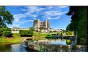 A large multi-story building set in a landscaped area with a pond in the foreground, surrounded by green trees and grass under a blue sky with some clouds.