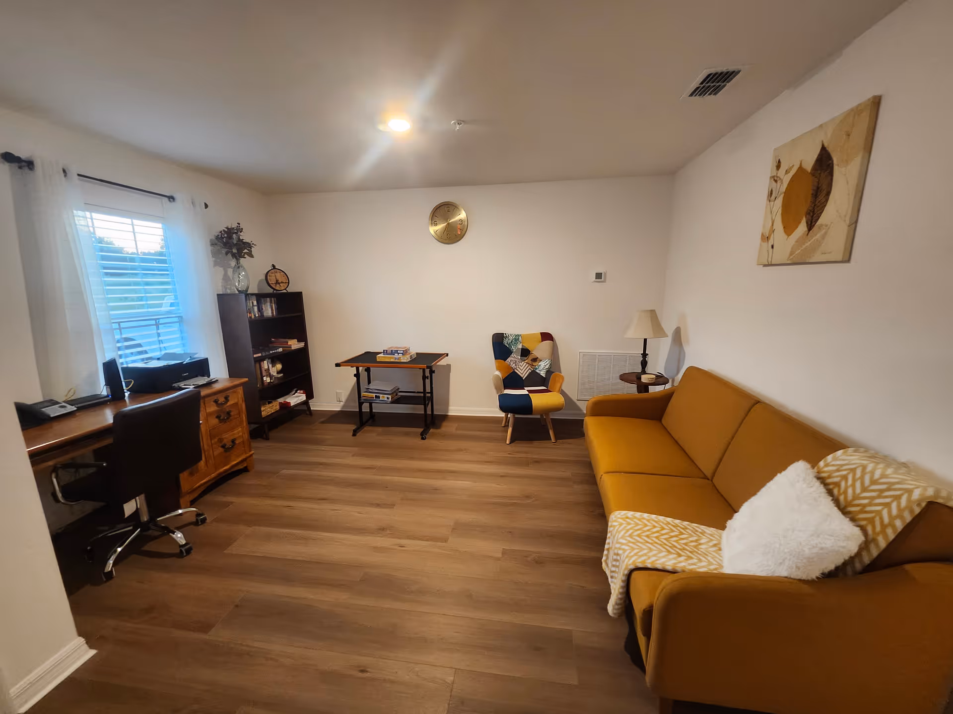 Bright living room with a mustard sofa, colorful armchair, desk, bookshelf and window on wood flooring.
