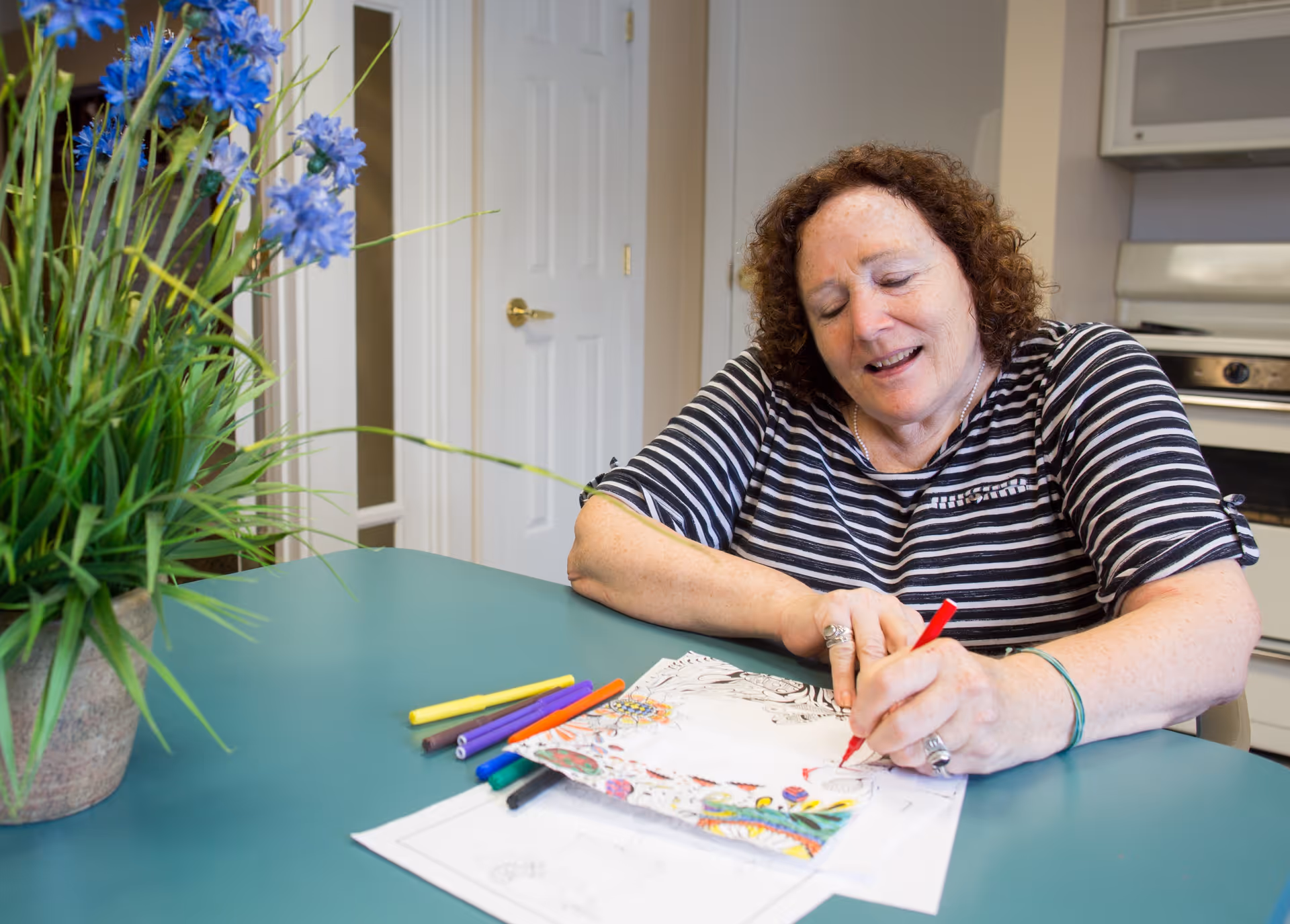 A woman with curly hair wearing a striped shirt is sitting at a teal table, coloring a detailed coloring page with a red marker. Several other colored markers are scattered on the table. There is a potted plant with blue flowers on the left side of the table, and a white door and kitchen appliances are visible in the background.