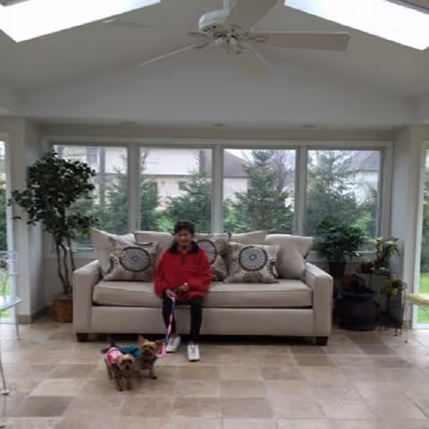 A woman in a red jacket sitting on a beige sofa with patterned cushions in a bright room with large windows and plants. Two small dogs on leashes are standing on the tiled floor in front of her.